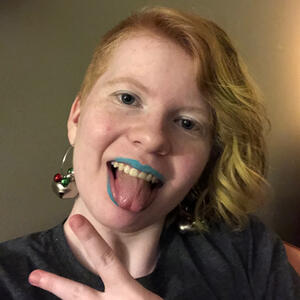 A selfie of a white teenager with loose green curls sticking their tongue out. They have light blue lipstick and large earrings.
